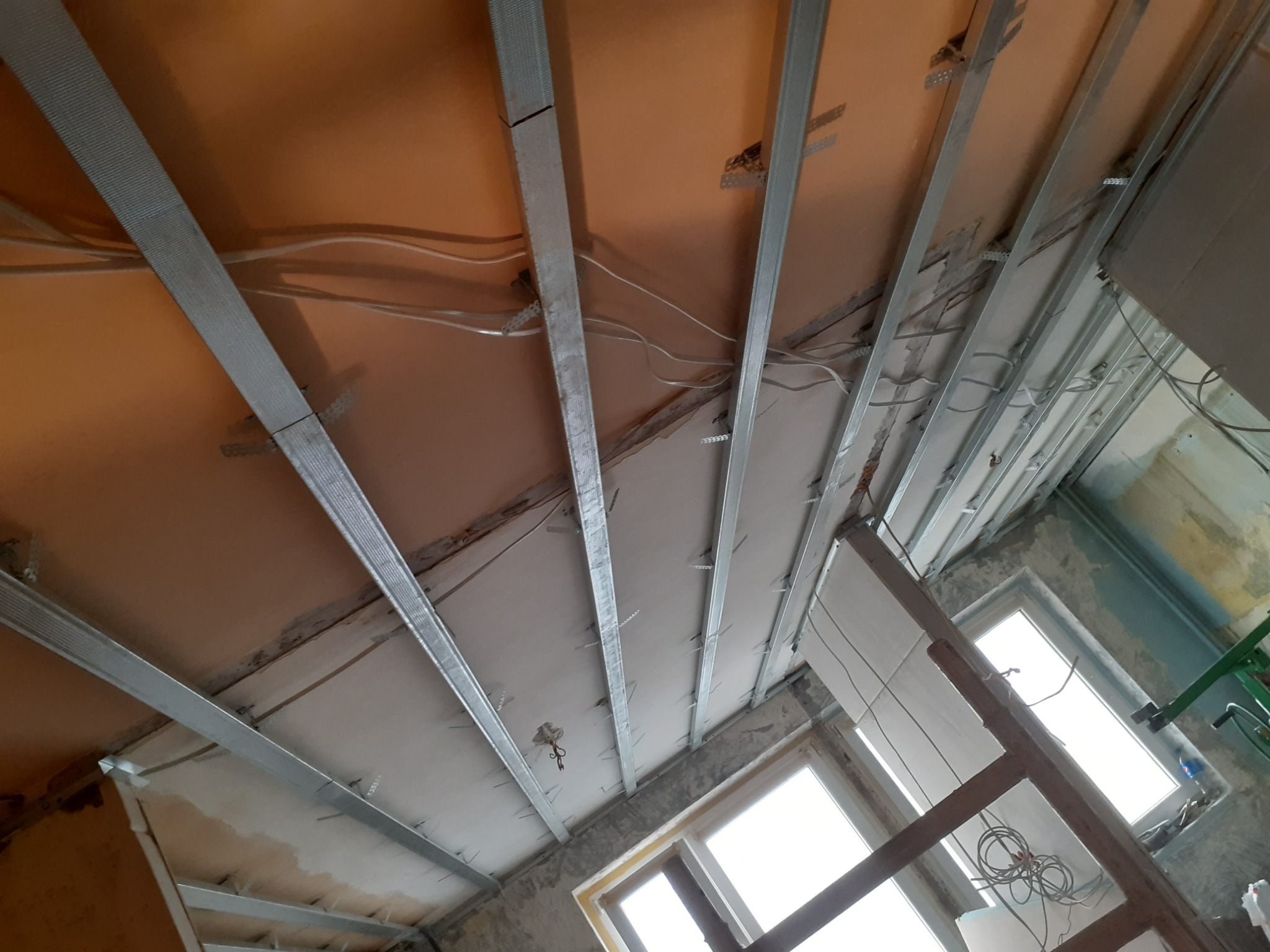 Widok z dołu na metalowy stelaż podwieszanego sufitu z płyt kartonowo-gipsowych, widoczne kable i częściowo zamontowane płyty przy oknach, surowy stan wykończenia.