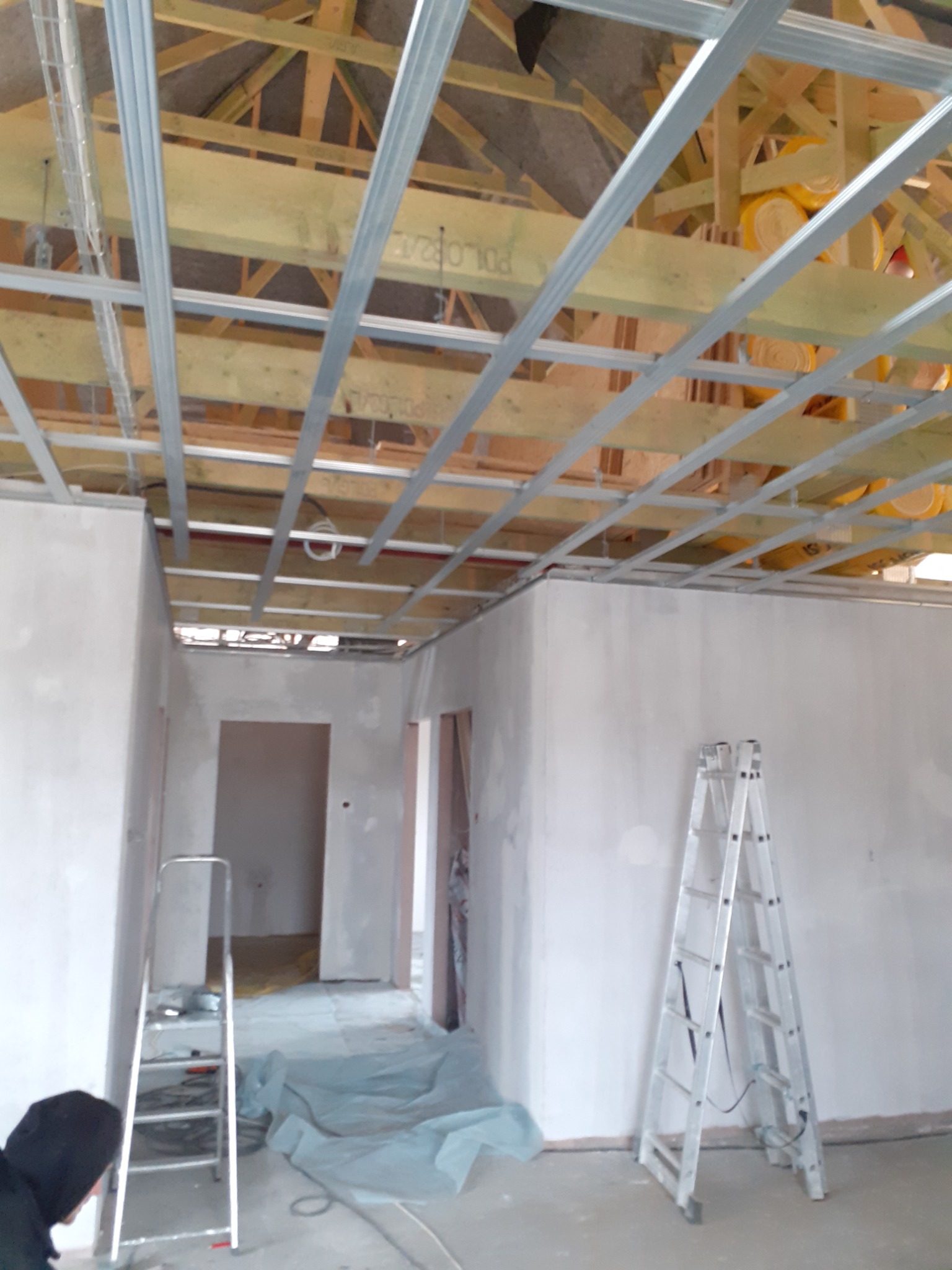 Montaż metalowej konstrukcji sufitu podwieszanego z widocznymi elementami izolacji termicznej, dwie drabiny aluminiowe w surowym wnętrzu budynku w trakcie prac wykończeniowych.