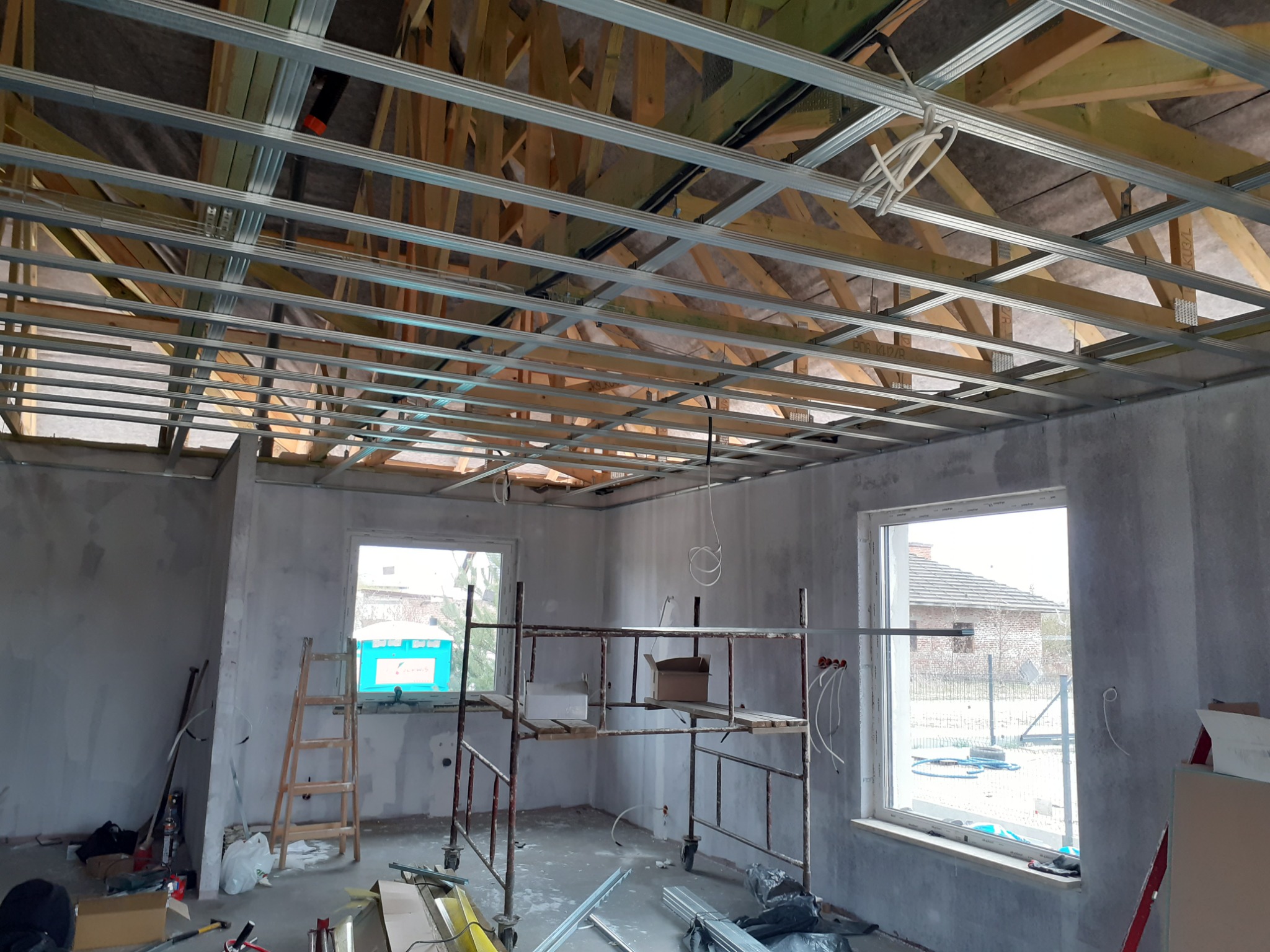Wnętrze w trakcie remontu: zamontowana metalowa konstrukcja sufitu podwieszanego z widocznymi elementami drewnianymi, surowe ściany, rusztowanie i drabina w pomieszczeniu.