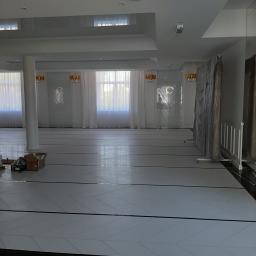 STAN-BUD - Sala balowa w trakcie prac wykończeniowych: podłoga z białych płytek ułożonych w jodełkę, kartony z materiałami budowlanymi w rogu, zabezpieczone folią elementy dekoracyjne.