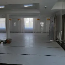 Sala balowa w trakcie prac wykończeniowych: podłoga z białych płytek ułożonych w jodełkę, kartony z materiałami budowlanymi w rogu, zabezpieczone folią elementy dekoracyjne.