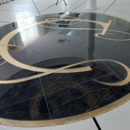 STAN-BUD - Okrągła posadzka z czarnego marmuru z intarsjami z metalu w kolorze złotym, tworzącymi abstrakcyjny wzór przypominający kaligrafię, otoczona białymi płytkami podłogowymi.