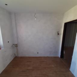 STAN-BUD - Pokój po malowaniu ścian na szaro, z drewnianą podłogą, widoczne puszki i kable elektryczne, instalacja wodna i materiały budowlane przy oknie.