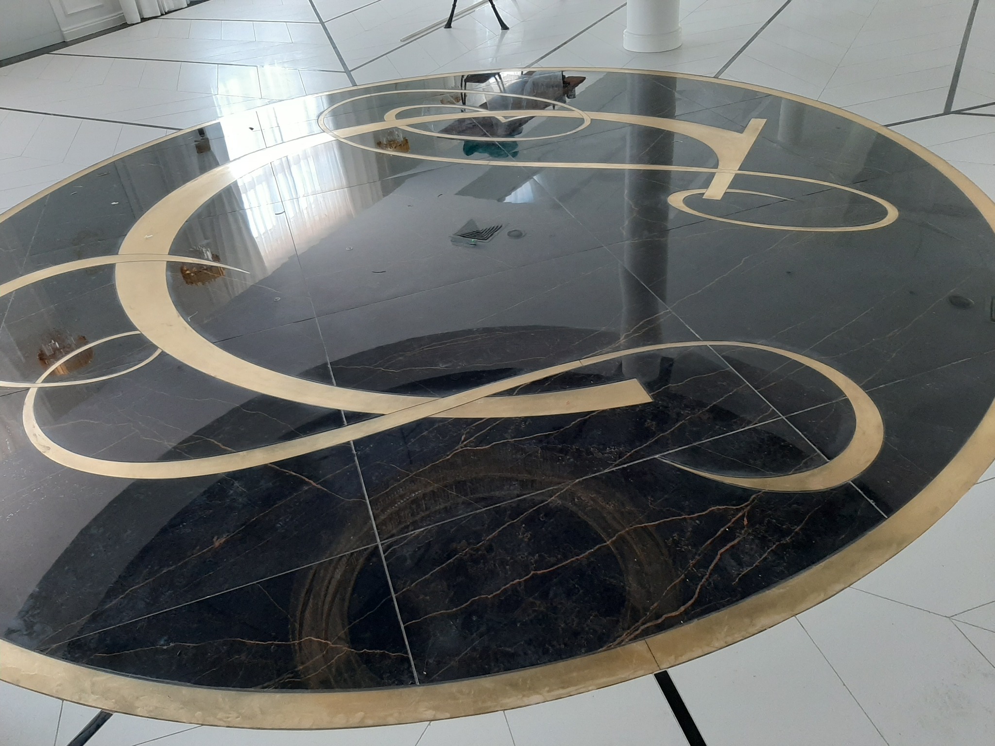 Okrągła posadzka z czarnego marmuru z intarsjami z metalu w kolorze złotym, tworzącymi abstrakcyjny wzór przypominający kaligrafię, otoczona białymi płytkami podłogowymi.