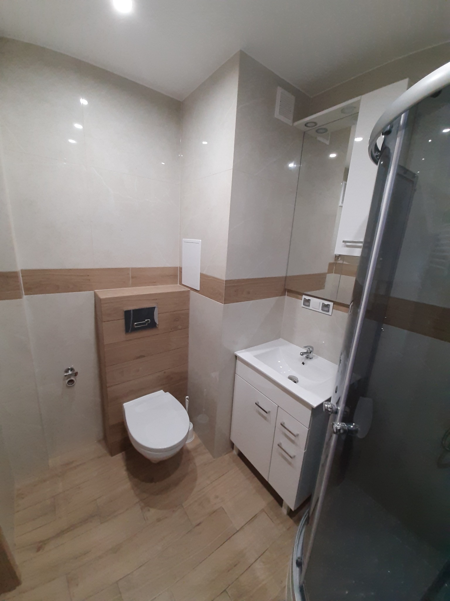 Nowoczesna łazienka z beżowymi płytkami, drewnianymi akcentami i białymi meblami: umywalka z szafką, toaleta podwieszana z drewnianą obudową, kabina prysznicowa z widocznym fragmentem ramy.