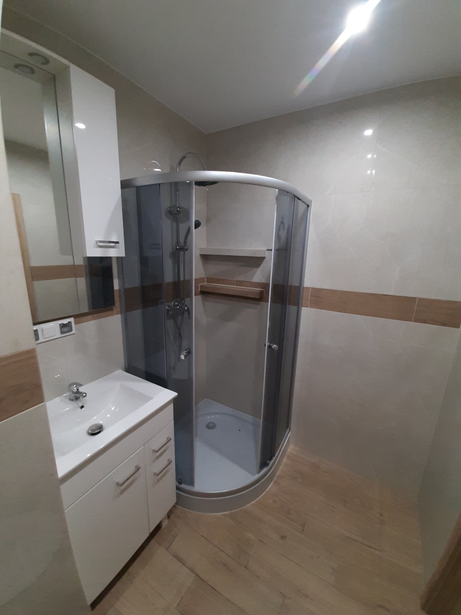 Nowoczesna łazienka z narożną kabiną prysznicową z szarymi szybami, białą szafką pod umywalką i lustrem, wykończona płytkami w odcieniach beżu z poziomym pasem dekoracyjnym.