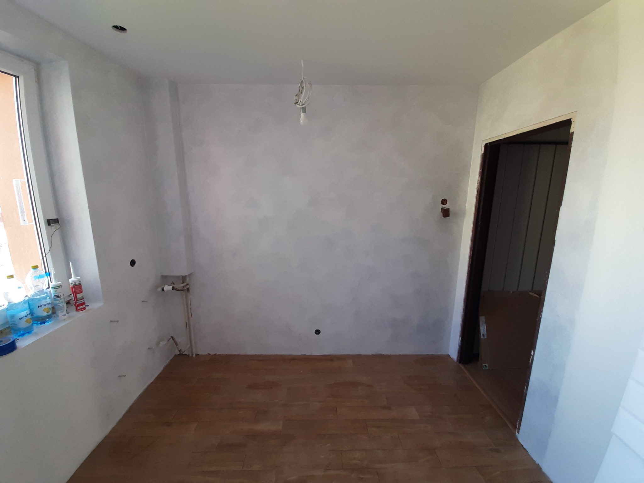 Pokój po malowaniu ścian na szaro, z drewnianą podłogą, widoczne puszki i kable elektryczne, instalacja wodna i materiały budowlane przy oknie.