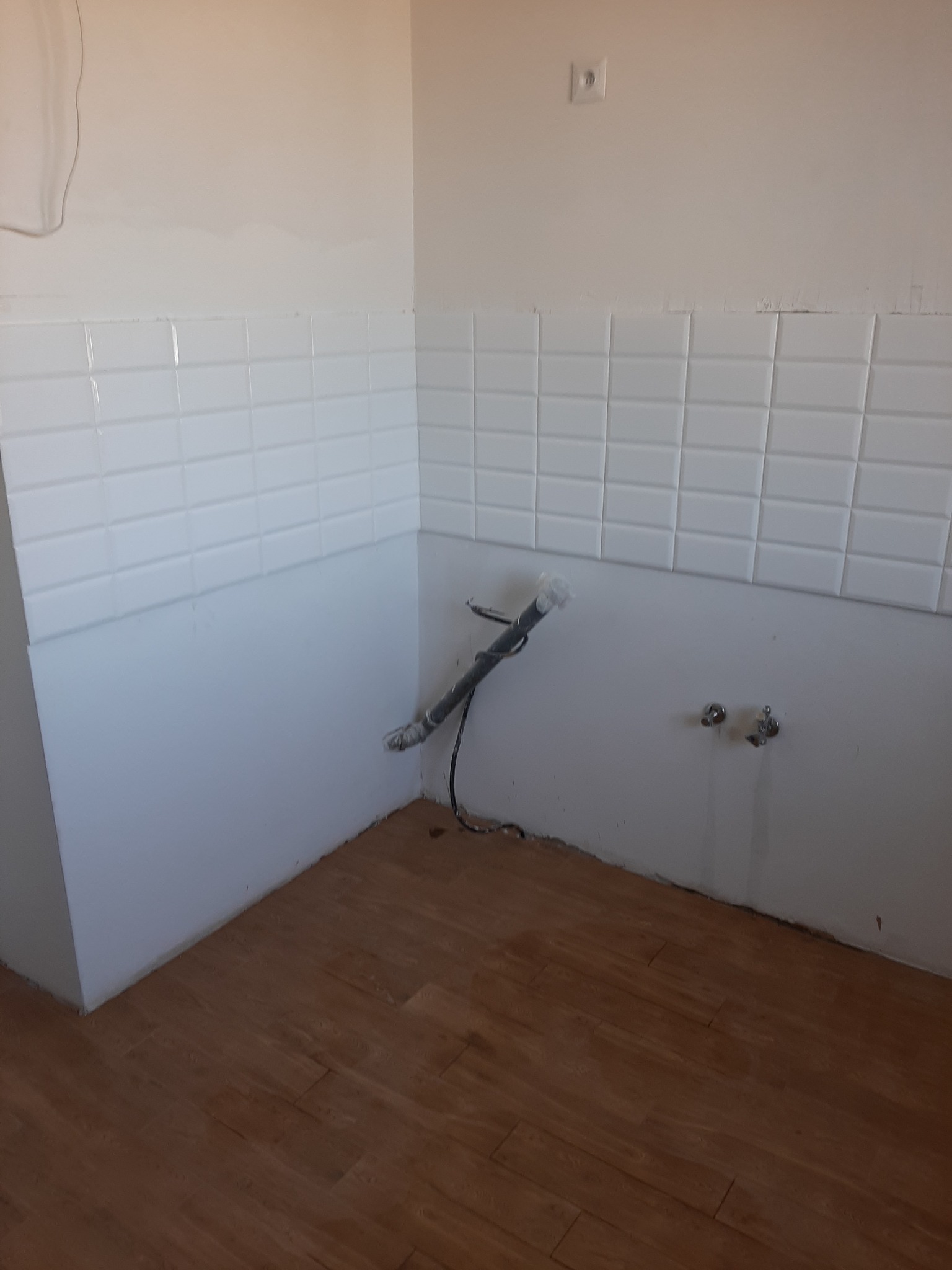 Fragment łazienki w trakcie prac: ułożone białe kafelki na ścianie do połowy wysokości, widoczne rury i zawory, brązowa podłoga imitująca drewno, ślady po remoncie na ścianie.