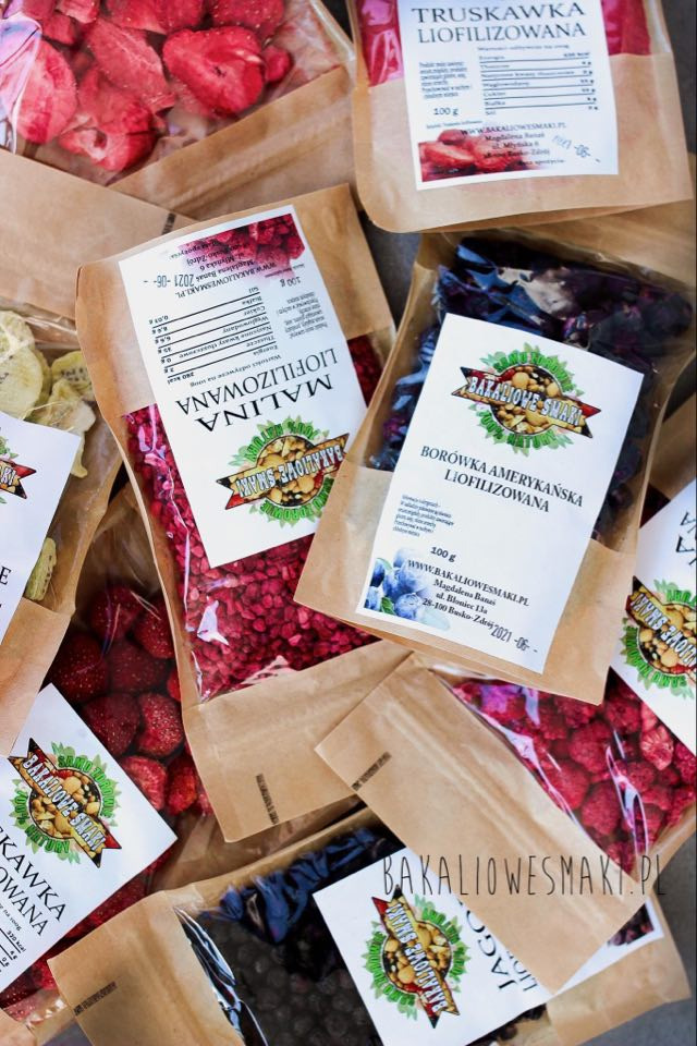 Różne rodzaje liofilizowanych owoców, takie jak truskawki, maliny i borówki amerykańskie, zapakowane w przezroczyste woreczki z etykietami 'Bakaliowe Smaki' na brązowym papierowym tle.