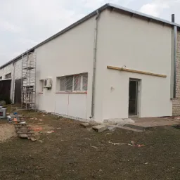 Budynek po ociepleniu i pomalowaniu elewacji na biało, z widocznymi zabezpieczonymi oknami folią, z resztkami materiałów budowlanych na trawniku i drabina rusztowaniowa z boku