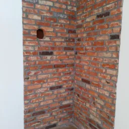 Fragment ściany wykończonej czerwoną cegłą klinkierową z widocznym otworem na instalację elektryczną, połączona z białą ścianą i podłogą imitującą drewno.