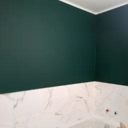Świeżo pomalowana ściana w łazience w kolorze butelkowej zieleni, kontrastująca z białymi marmurowymi płytkami i białą wanną wypełnioną wodą, widoczna drabina malarska.