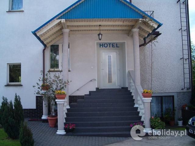 Wejście do hotelu z niebieskim daszkiem, schodami i kolumnami, z napisem HOTEL nad drzwiami.