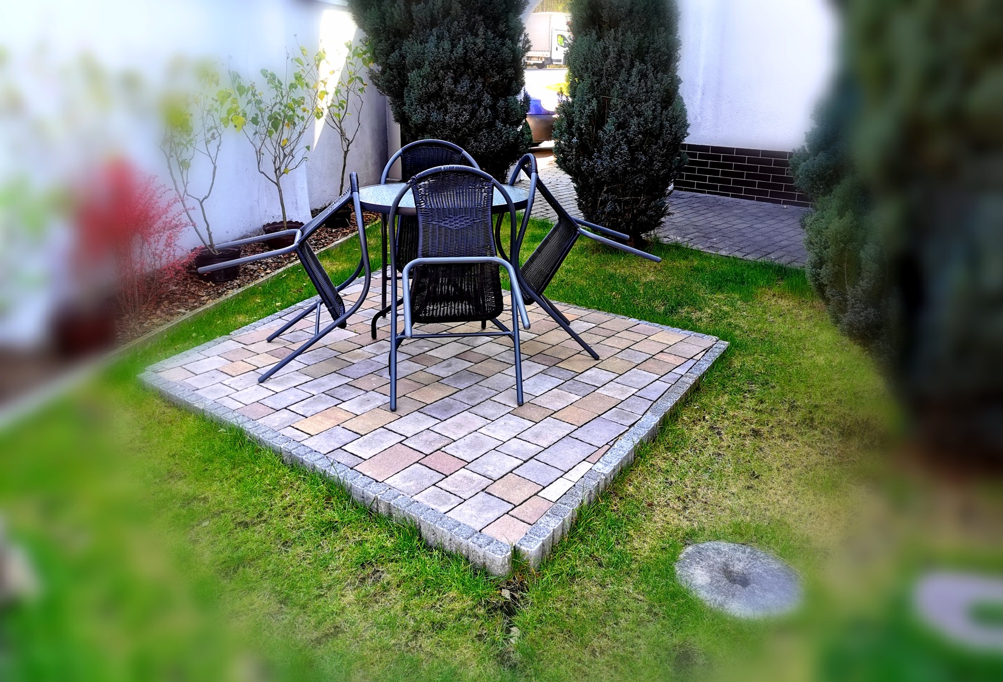 Zestaw mebli ogrodowych z plecionymi krzesłami i szklanym stołem na kwadratowym patio z kostki brukowej, otoczony trawnikiem i roślinnością.