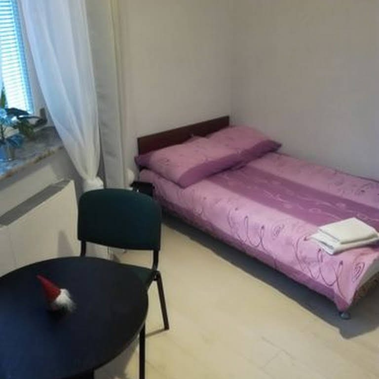 Pokój z łóżkiem pojedynczym w różowej pościeli z fioletowymi wzorami, mały stolik z czarnym blatem i dekoracyjnym krasnalem, krzesło, okno z zasłoną i parapetem z kwiatami.