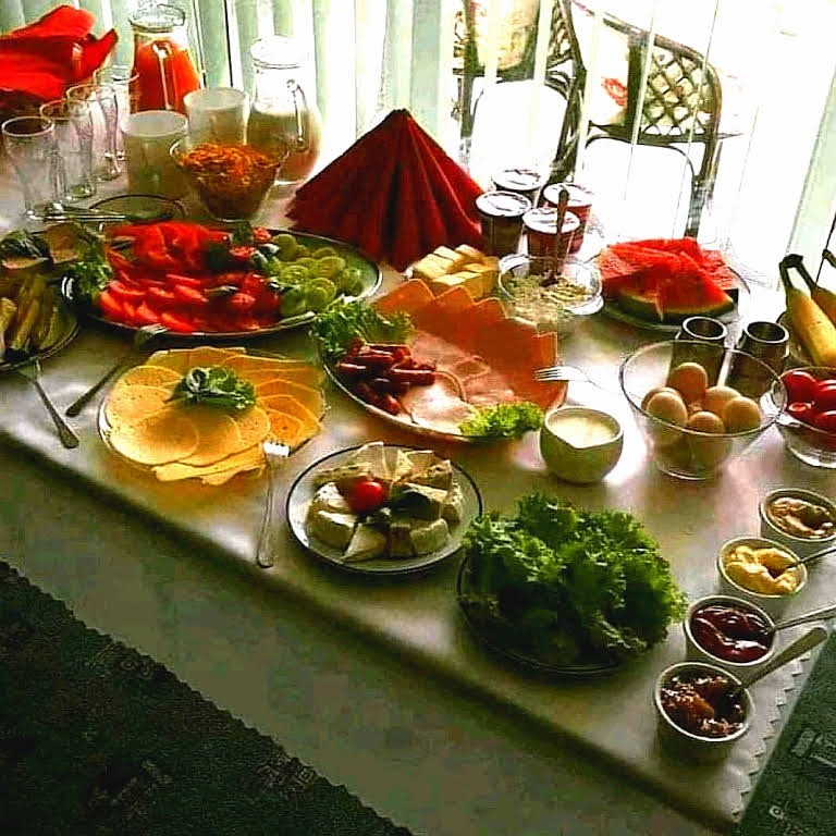 Stół bufetowy z różnorodnym śniadaniem: wędliny, sery, warzywa, owoce, jajka, dżemy i napoje w szklanych dzbankach, ustawione przy oknie z białymi zasłonami.