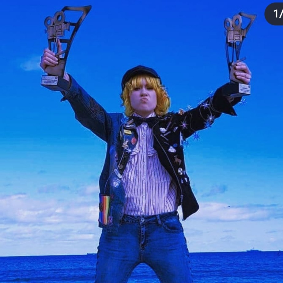 Osoba z blond włosami, w kapeluszu i ekstrawaganckiej marynarce, trzymająca w górze dwie statuetki w kształcie kamery filmowej na tle błękitnego nieba i morza.