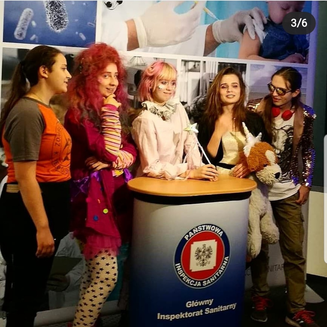 Grupa młodych osób w kostiumach stoi przy stoisku Państwowej Inspekcji Sanitarnej, na tle zdjęć mikroskopowych i szczepień dzieci.
