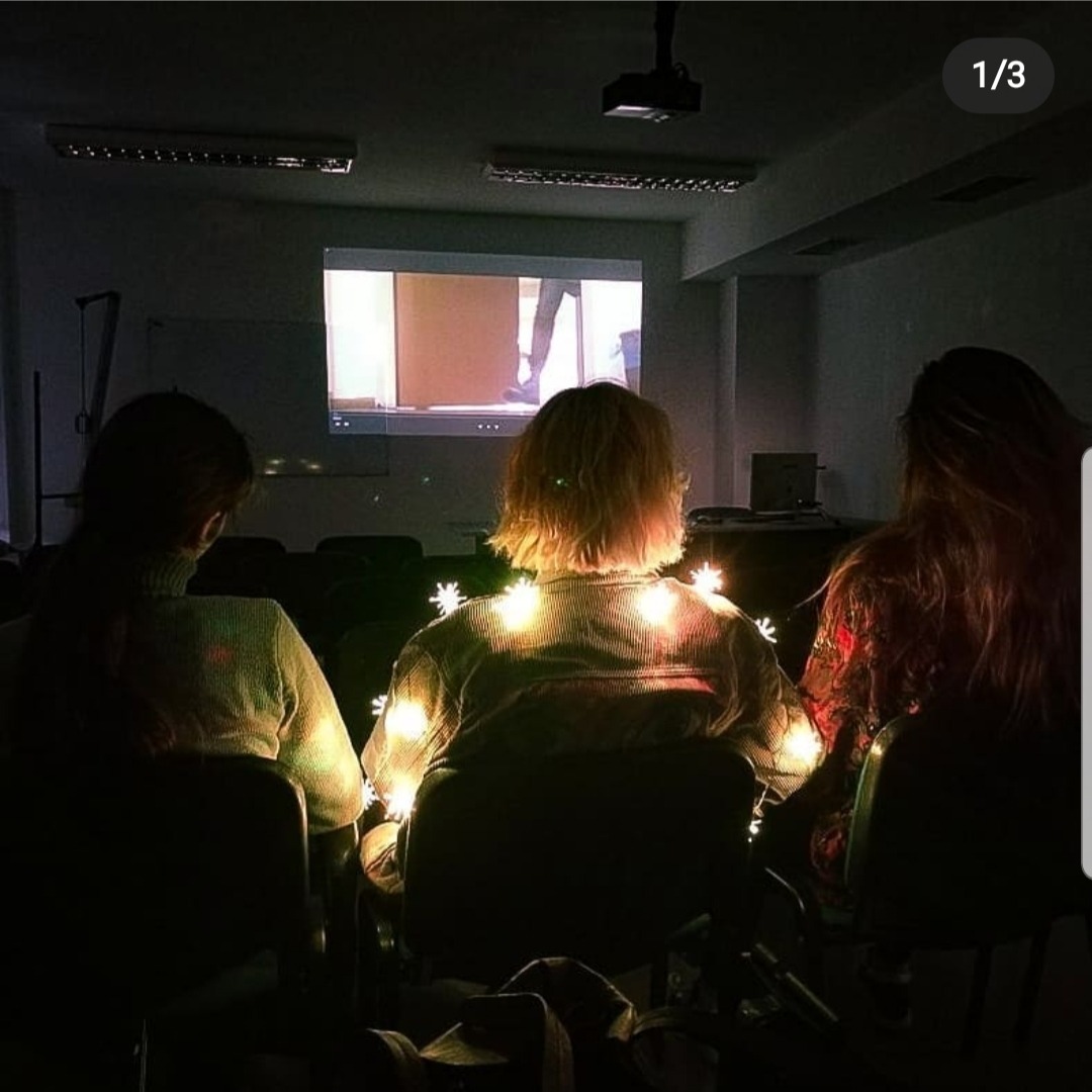 Trzy osoby siedzą w ciemnym pomieszczeniu, oglądając film wyświetlany na ekranie projektora. Świąteczne lampki owinięte wokół środkowej osoby dodają ciepłego blasku.