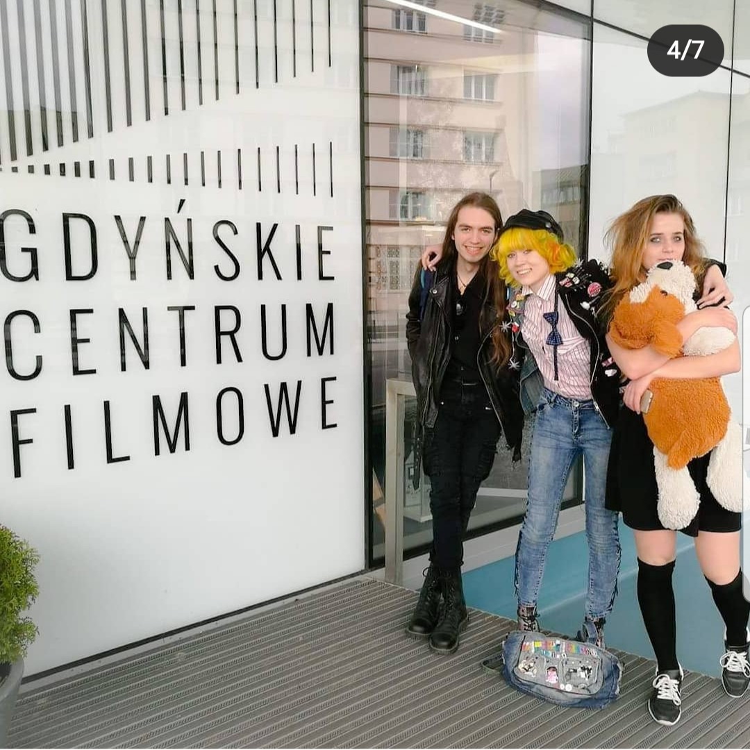 Trzy osoby pozują przed Gdyńskim Centrum Filmowym, jedna z nich trzyma pluszowego misia, widoczny plecak z przypinkami.