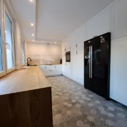 Nowoczesna kuchnia z białymi szafkami, drewnianym blatem, czarną armaturą i lodówką oraz heksagonalną podłogą w odcieniach szarości.