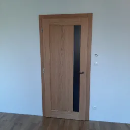 Jasne drewniane drzwi wewnętrzne z pionowym czarnym paskiem, zamontowane w białej ścianie, widoczna podłoga z paneli.