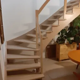 Drewniane schody z jasnego drewna z szklaną balustradą, widok z dołu, w tle rośliny doniczkowe i fragment pomarańczowego stołu.