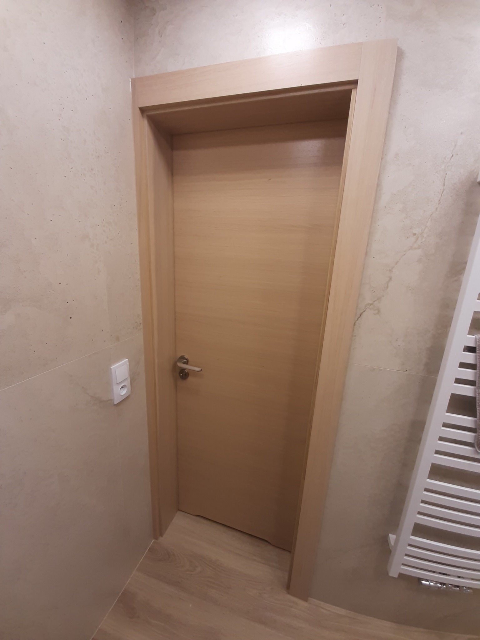 Jasne, drewniane drzwi z prostą ościeżnicą w łazience z beżowymi płytkami i grzejnikiem drabinkowym.