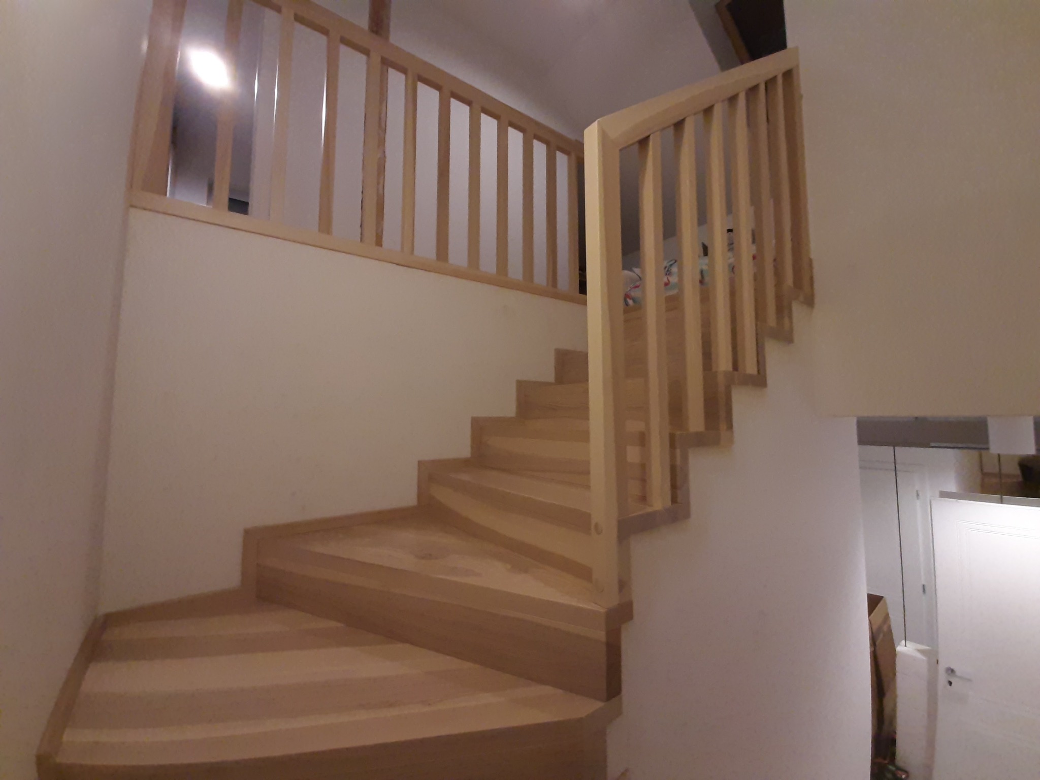 Jasne, drewniane schody z barierką, prowadzące na antresolę w nowoczesnym wnętrzu. Widoczne proste, pionowe szczebelki barierki i delikatny skręt schodów.