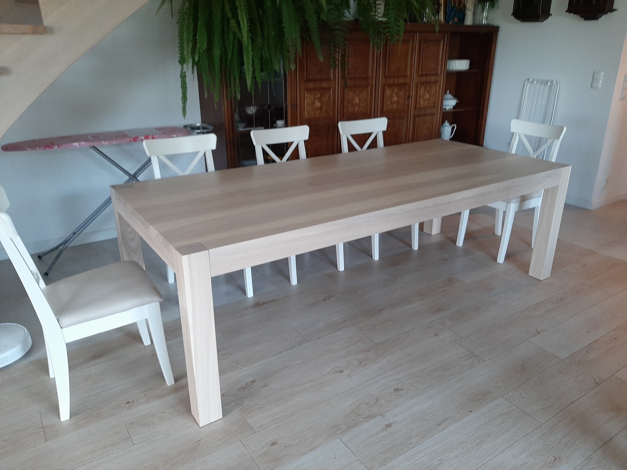 Jasny, drewniany stół jadalny z białymi krzesłami w minimalistycznym wnętrzu, z widoczną deską do prasowania i szafką z dekoracjami w tle.