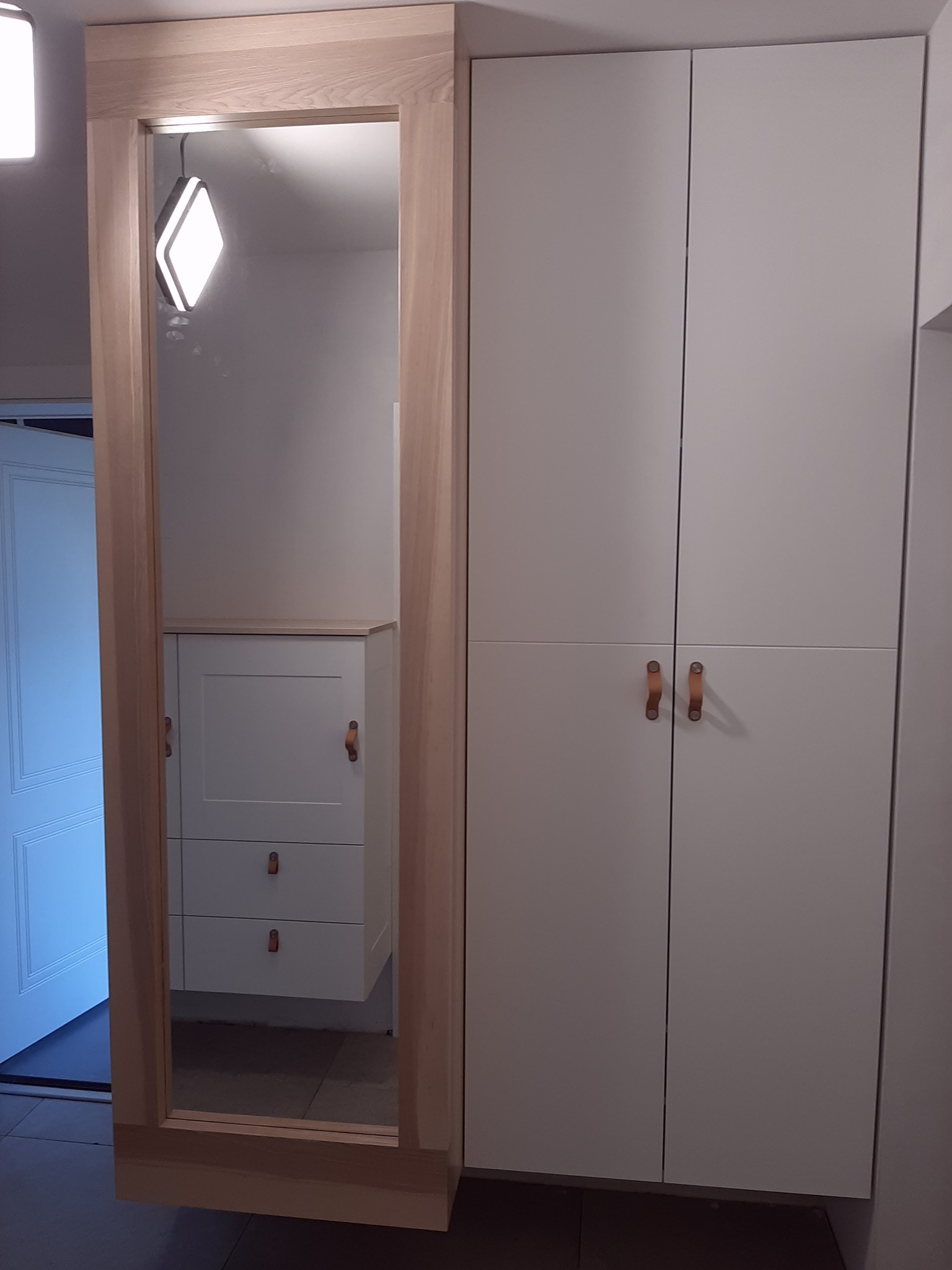 Biała szafa z drewnianymi uchwytami, obok lustro w drewnianej ramie odbijające białą komodę z szufladami i oświetlenie.