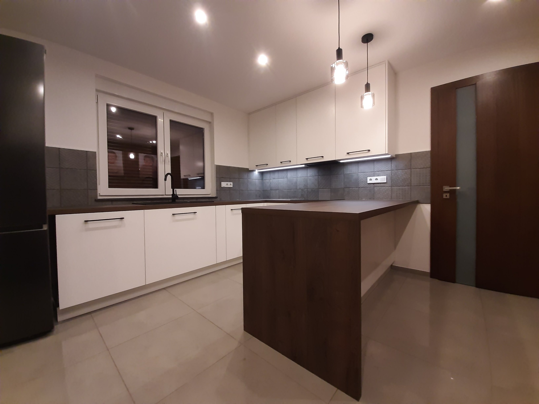 Nowoczesna kuchnia z białymi szafkami, ciemnym blatem i wyspą, oświetlona wiszącymi lampami, z widoczną lodówką i drzwiami z przeszkleniem.