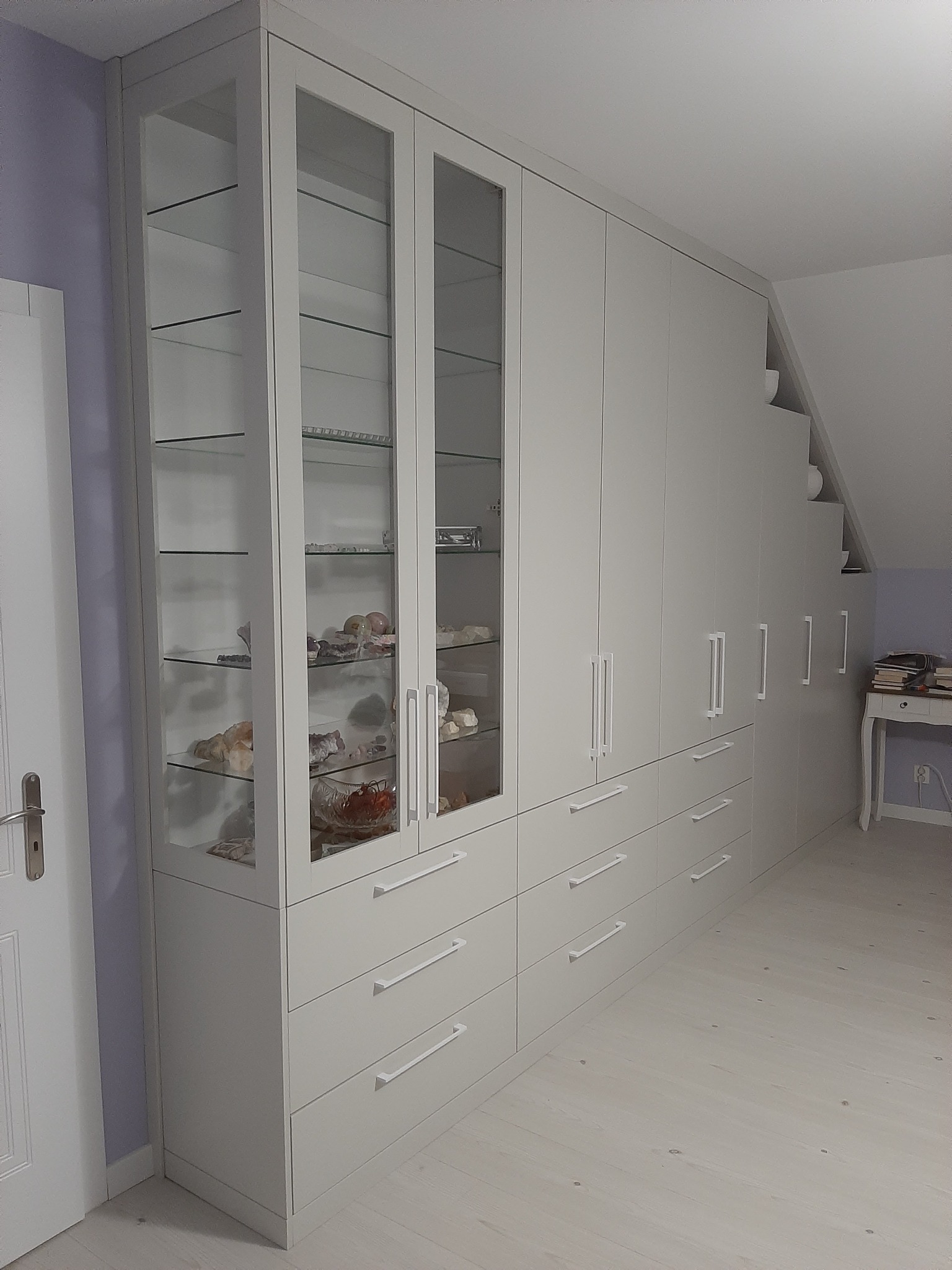 Jasnoszara zabudowa meblowa pod skosem, z przeszklonymi witrynami wypełnionymi kolekcją minerałów i kamieni, oraz rzędem szuflad z długimi uchwytami.