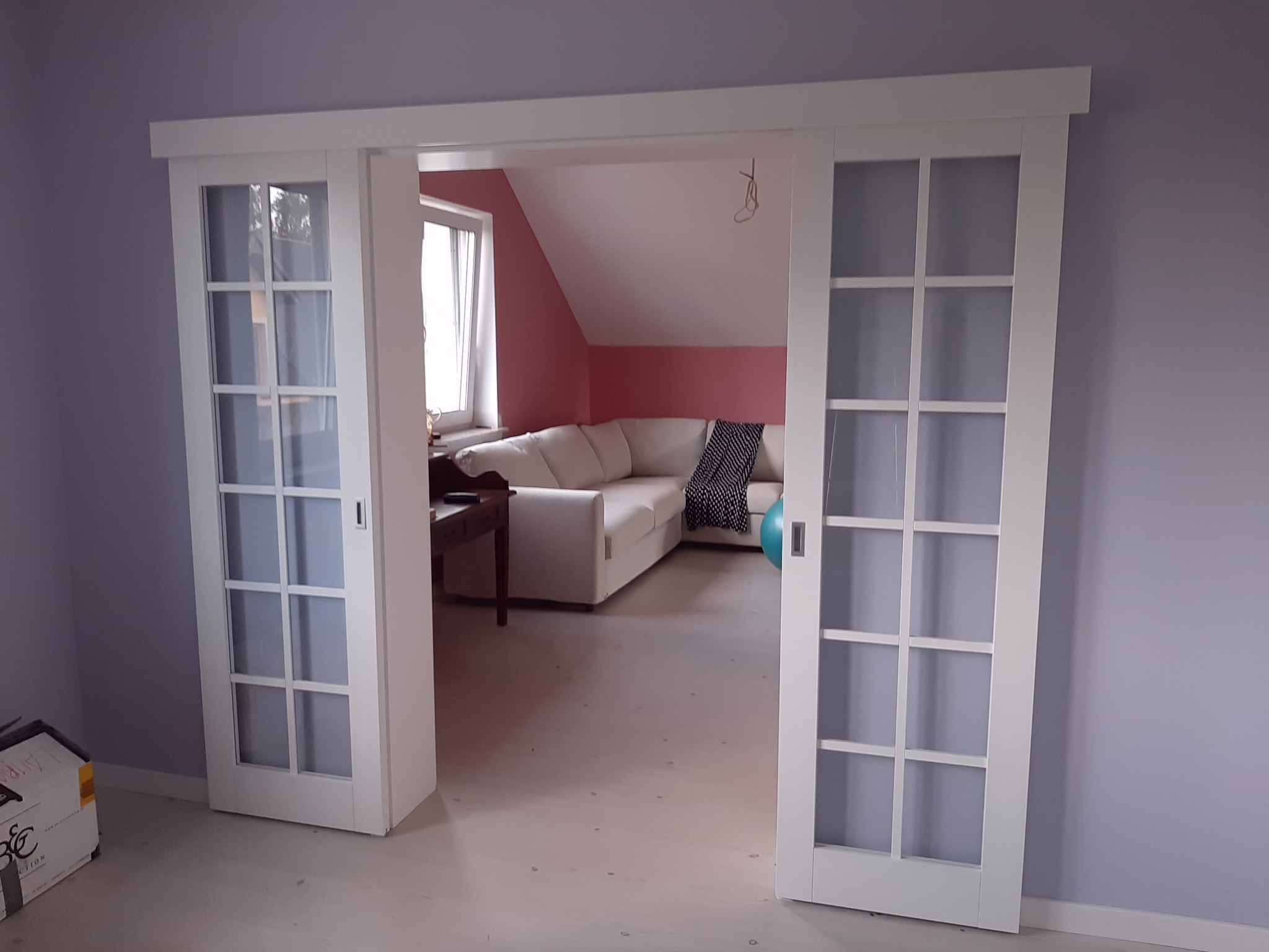 Białe drzwi przesuwne z szybami w ramkach, widok na salon z narożną sofą i skośnym sufitem, fioletowe ściany, karton na podłodze.