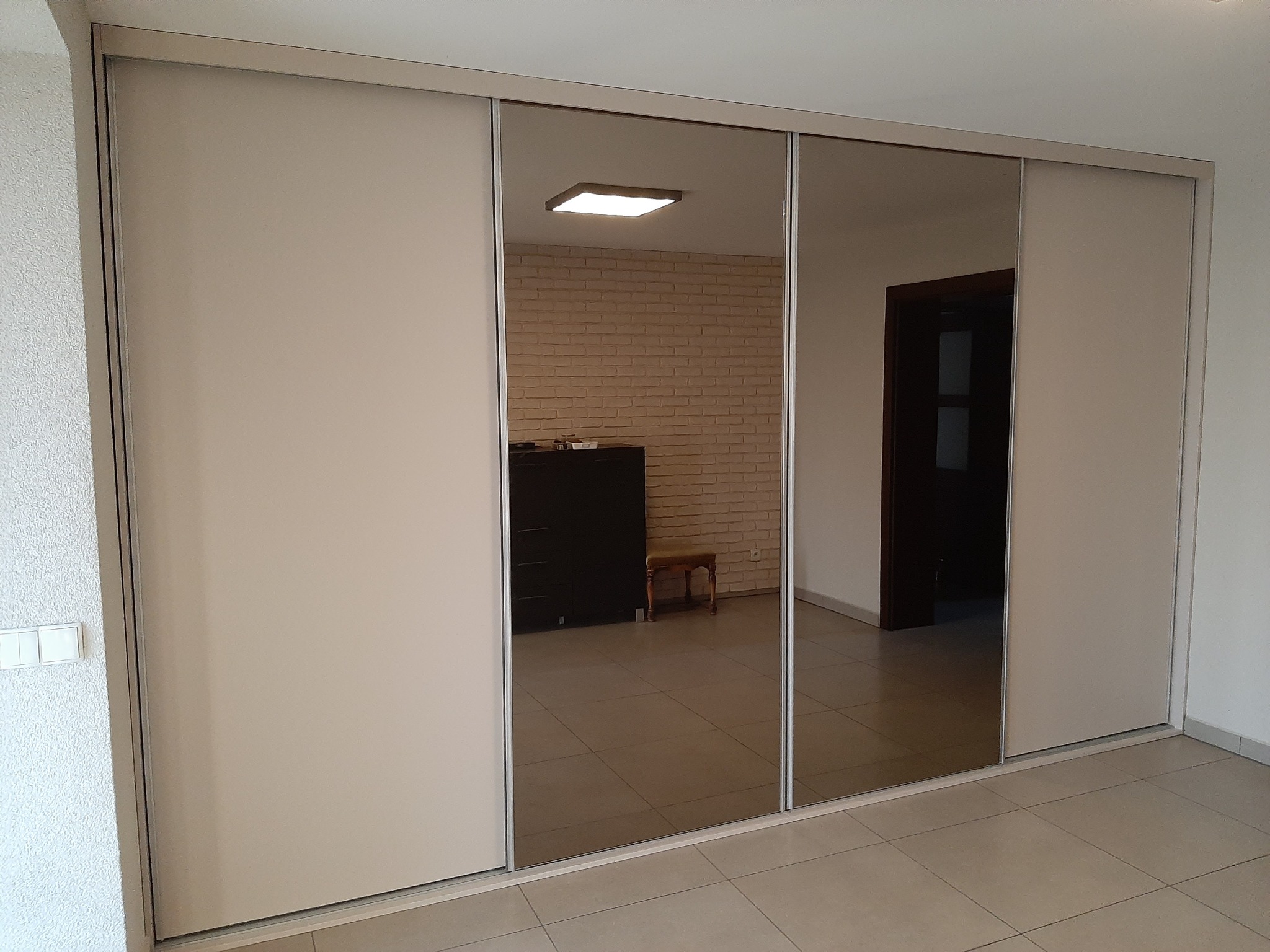 Beżowa szafa przesuwna z lustrzanymi drzwiami, odbijająca ceglaną ścianę i ciemny mebel w pokoju.