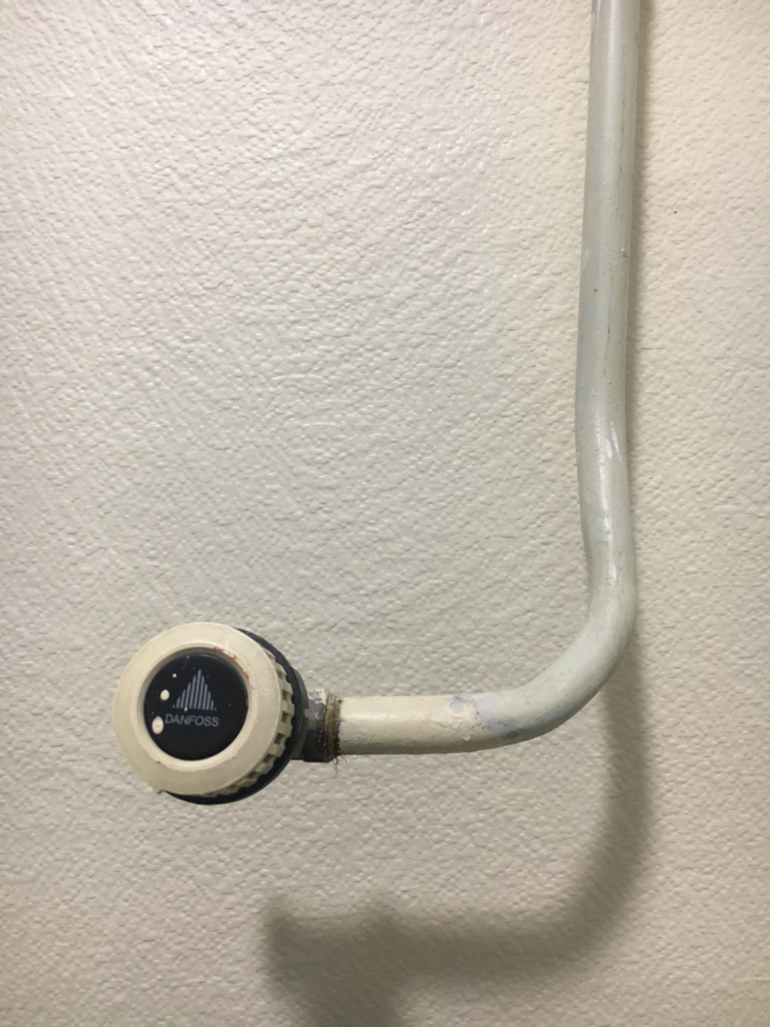 Biała, teksturowana ściana z widoczną, pomalowaną na biało rurą grzejnika i termostatem Danfoss. Widoczne ślady użytkowania i drobne niedomalowania na rurze i ścianie.