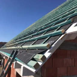 Konstrukcja dachu w trakcie budowy: widoczne drewniane łaty w kolorze turkusowym, czarna membrana dachowa i ceglany mur budynku, z drabiny opierającej się o krawędź dachu.