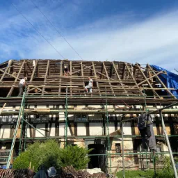 Demontaż starego dachu na budynku szachulcowym z rusztowaniem, widoczni pracownicy w trakcie pracy, część dachu zabezpieczona plandeką.