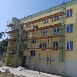 Żółto-zielony budynek w trakcie prac wykończeniowych, z rusztowaniem i pracownikami na różnych poziomach, widoczne materiały budowlane i narzędzia na ziemi.