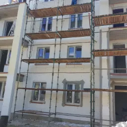 Wznoszenie ścian domu z bloczków betonu komórkowego Ytong, widoczne rusztowanie i pracownik montujący okno na wyższej kondygnacji.