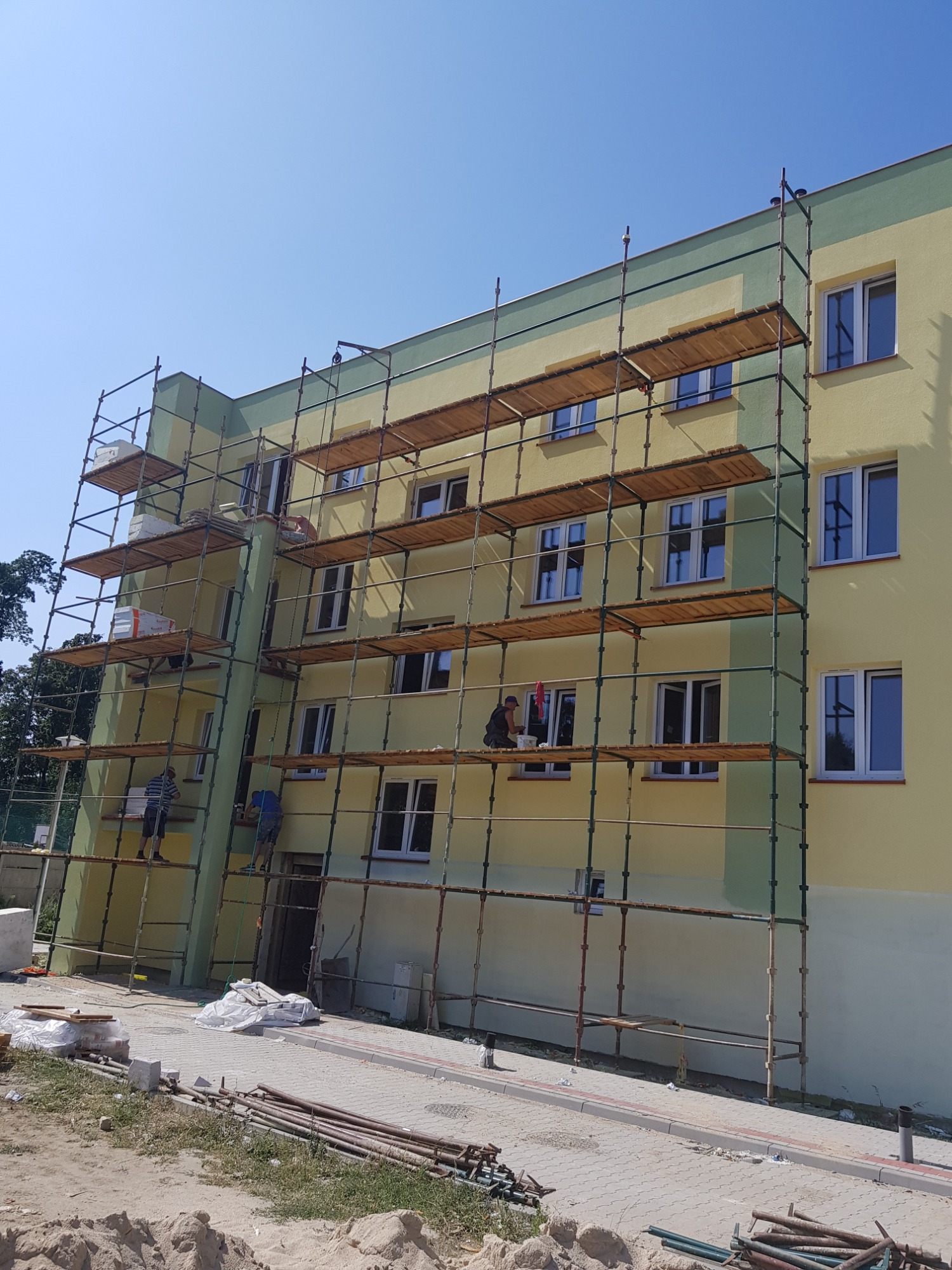 Żółto-zielony budynek w trakcie prac wykończeniowych, z rusztowaniem i pracownikami na różnych poziomach, widoczne materiały budowlane i narzędzia na ziemi.