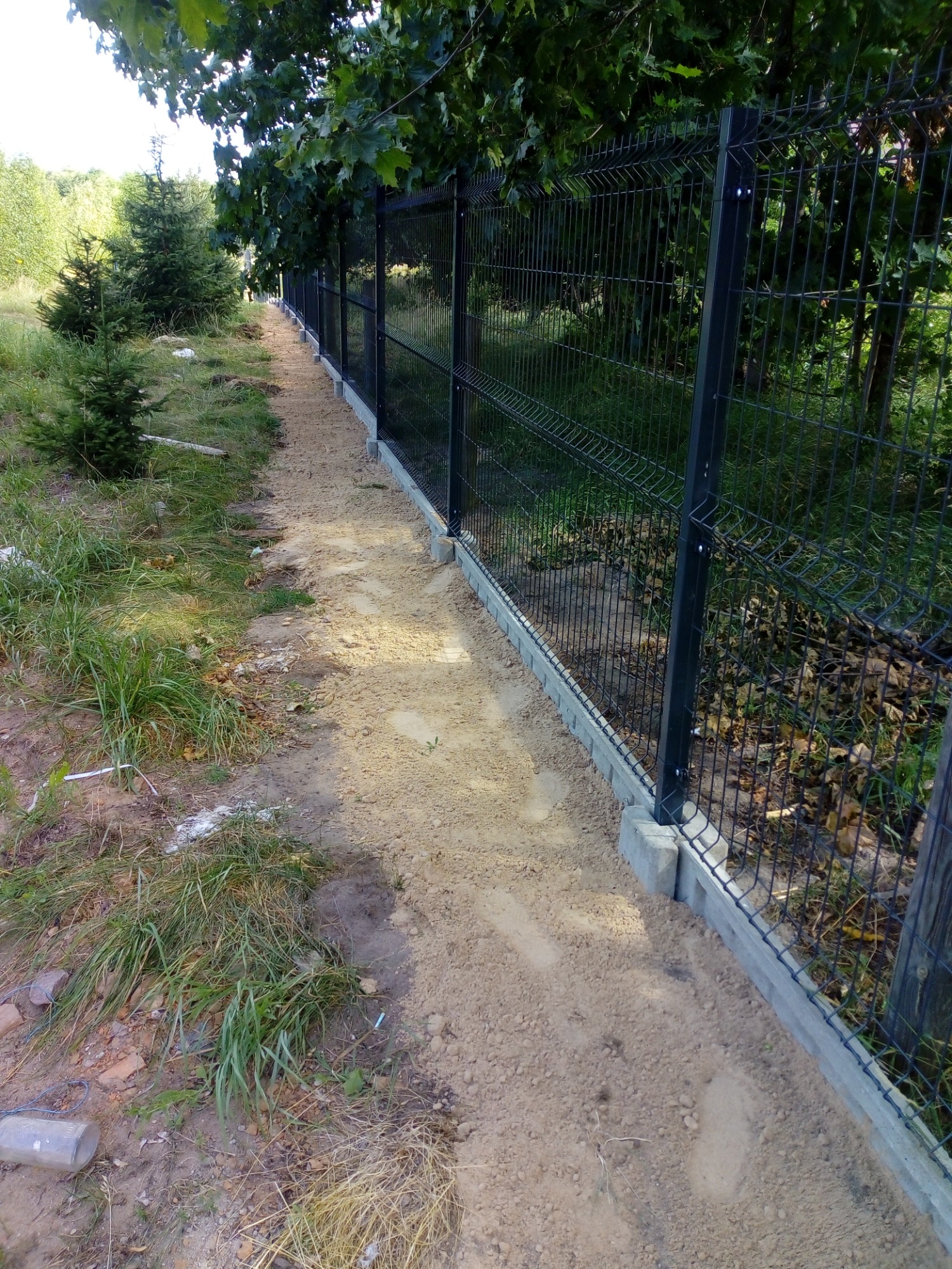 Długa linia czarnego ogrodzenia z siatki panelowej na betonowej podmurówce, widok z boku na nierówny teren z drzewami w tle.