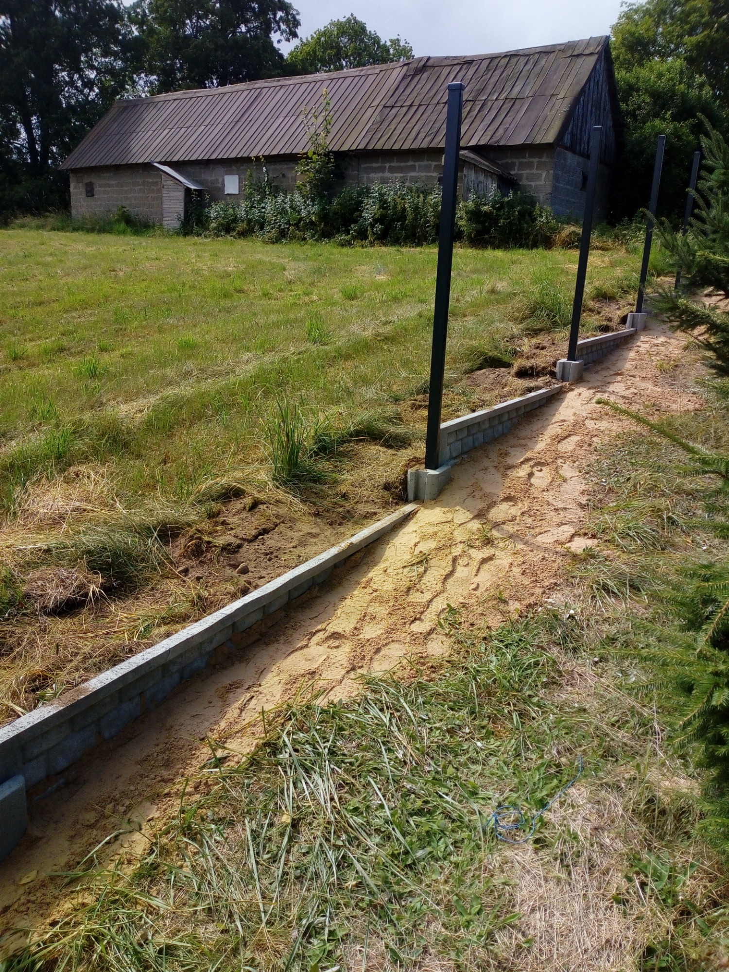 Wykop pod fundament ogrodzenia z wylaną podmurówką z betonowych bloczków i zamontowanymi słupkami, widok na wiejski krajobraz z budynkiem gospodarczym w tle.