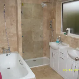 Wykończona łazienka z beżowymi płytkami, kabiną prysznicową ze szklanymi drzwiami, białą wanną i szafką z dwoma umywalkami, widok z otwartych drzwi.