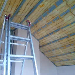 Montaż izolacji termicznej na suficie podwieszanym z metalowym stelażem, widoczna drabina aluminiowa.