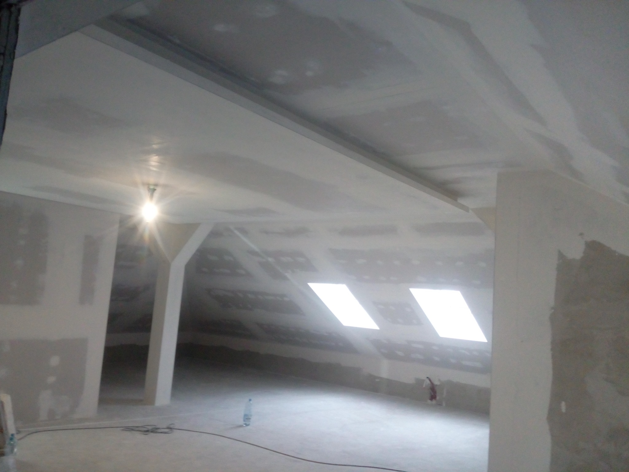 Poddasze w trakcie wykańczania, widoczne zamontowane profile pod konstrukcję sufitu, częściowo pokryte szpachlą, w tle okna dachowe.