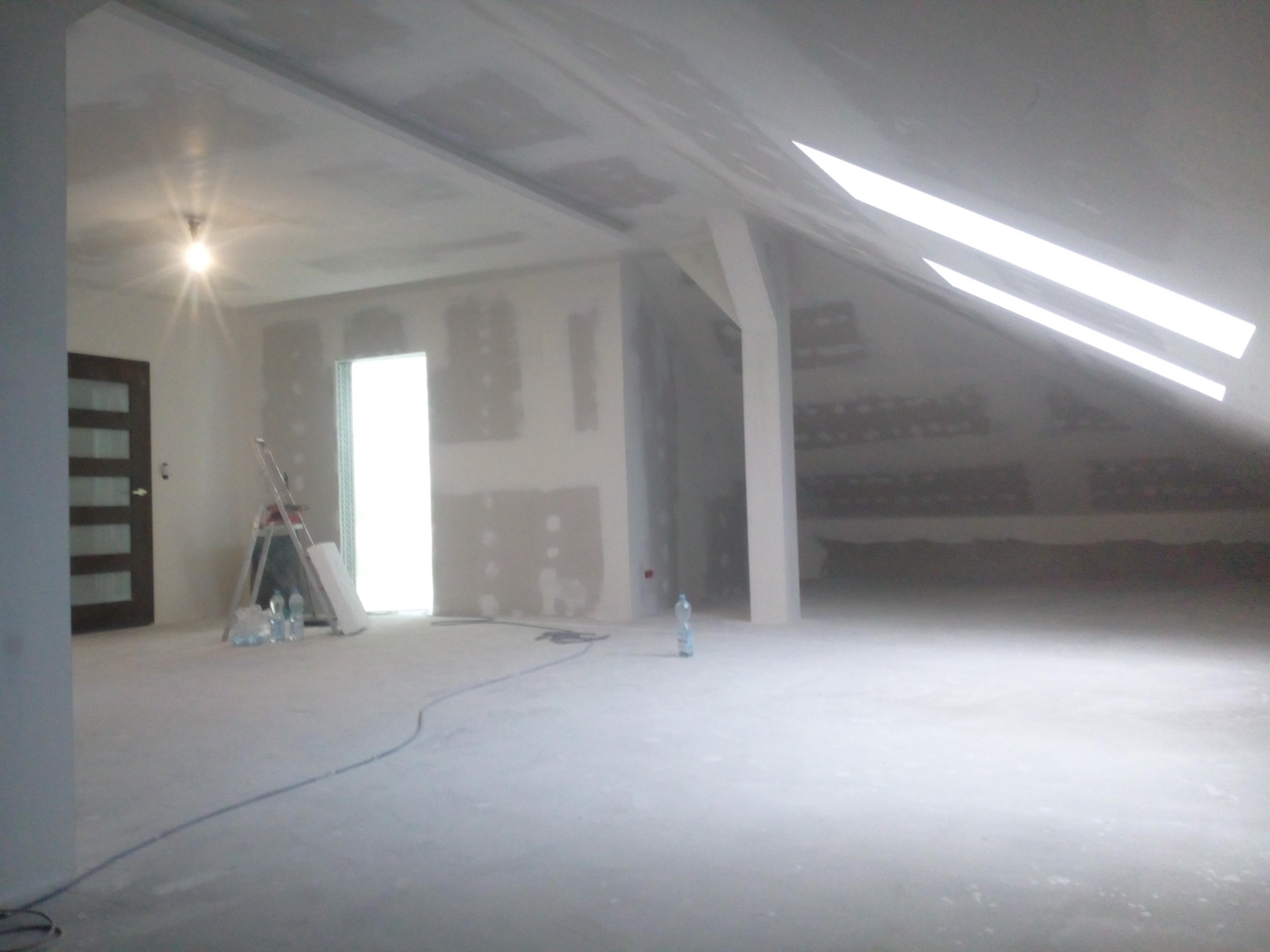 Poddasze w trakcie remontu, widoczne zamontowane profile pod płyty, częściowo położona gładź szpachlowa, rozstawiona drabina i butelki z wodą.