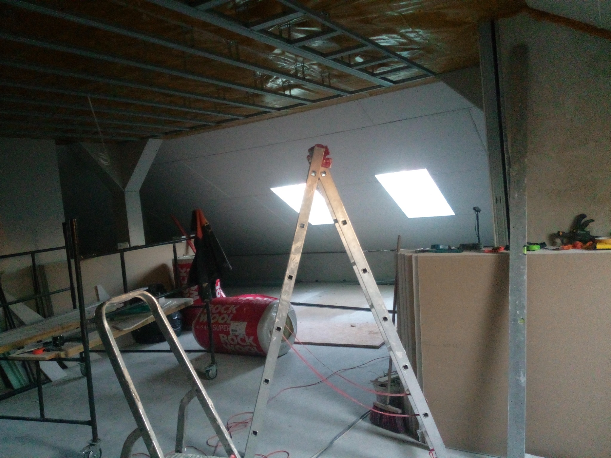 Montaż sufitu podwieszanego na poddaszu: widoczna konstrukcja metalowa, izolacja z wełny mineralnej, ustawiona drabina, przygotowane płyty oraz okna dachowe.