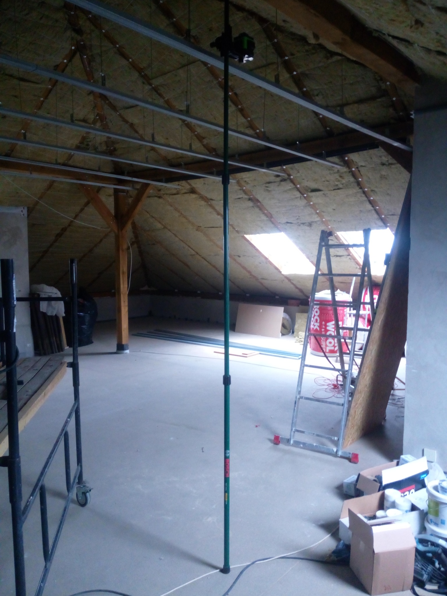 Poddasze w trakcie adaptacji: widoczne profile pod zabudowę, izolacja wełną mineralną, laser krzyżowy na statywie, drabina i materiały budowlane.