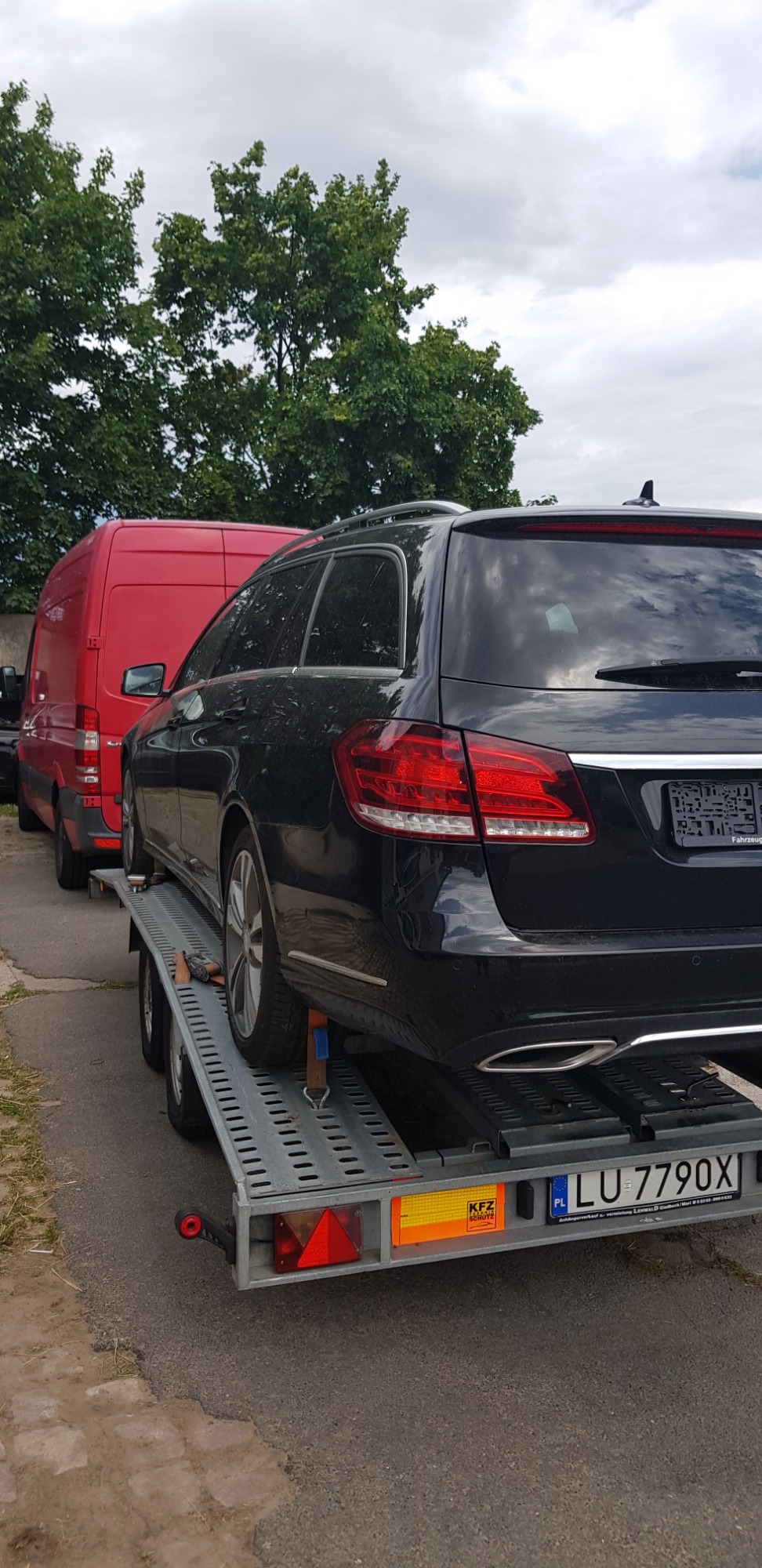 Czarny samochód kombi marki Mercedes-Benz na lawecie transportowej z polską tablicą rejestracyjną LU 7790X, widoczny czerwony samochód dostawczy w tle.